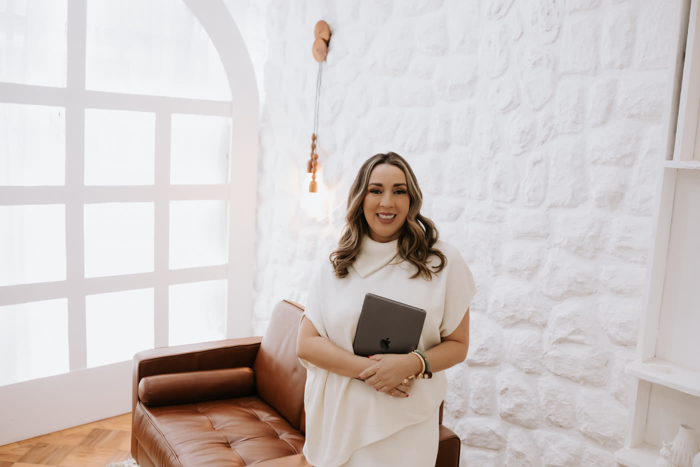 Edkarla Almeida em pé, com roupa creme, segurando um tablet em uma sala com arco e parede de pedra branca